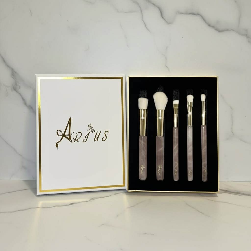 A 5-piece makeup brush set with rose quartz handles, gold ferrules, and white bristles, displayed in a black box next to its white lid which features the brand name 'Arius' in gold foil, set against a white marble background.